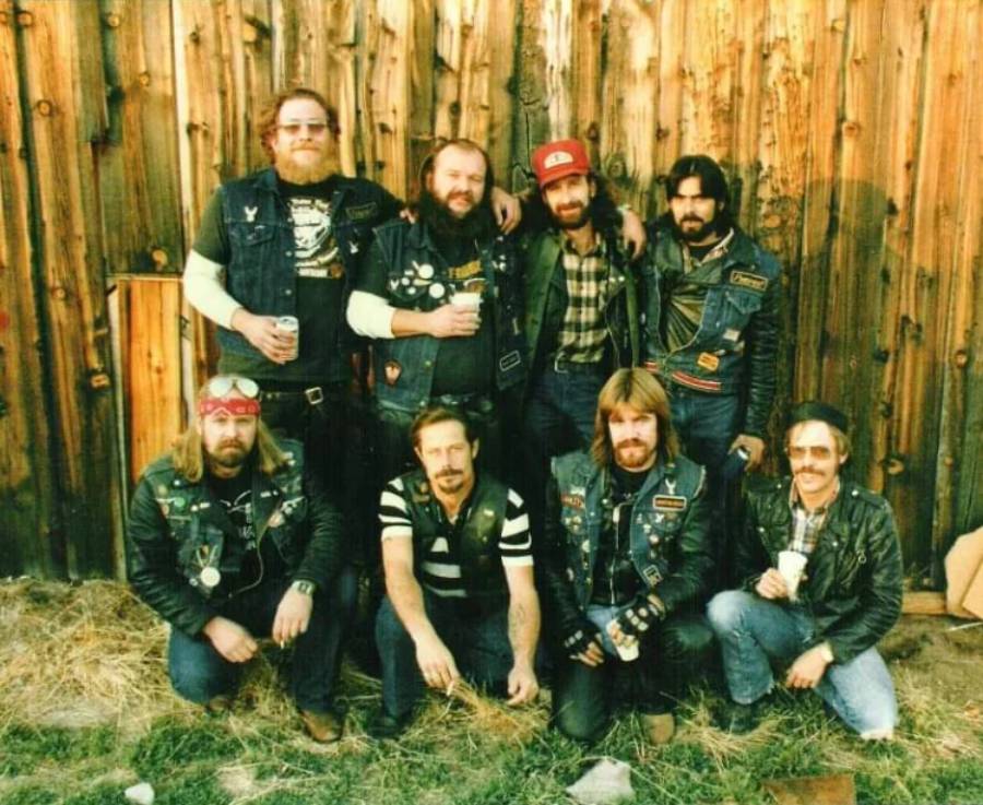 Eight men, some with beards and sunglasses, wearing denim or leather vests covered in patches, pose in front of a wooden wall outdoors. Most are standing, while three kneel or sit in front.