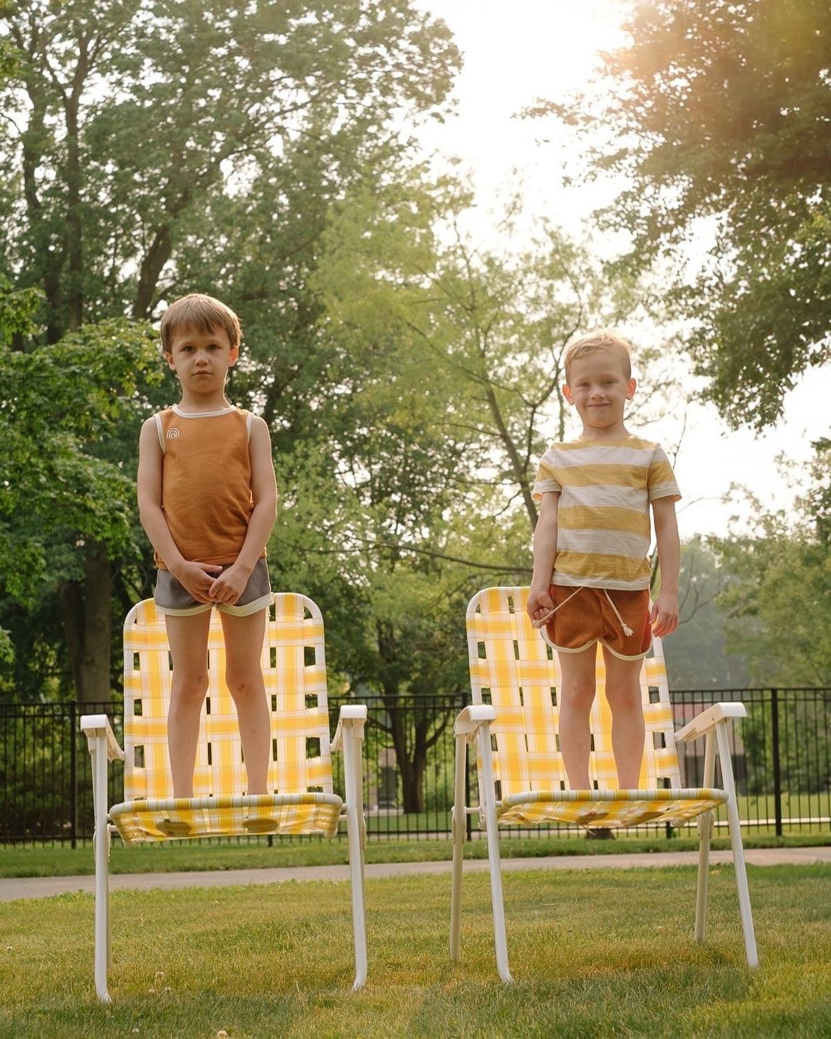 Two young children stand on yellow-patterned lawn chairs in a sunlit yard, wearing matching earth-tone outfits. Trees and a fence are visible in the background.