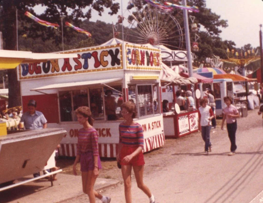 Three people walk past colorful fair food stands at a carnival, including one selling corn dogs. A Ferris wheel and other booths are visible in the background, with trees and flags overhead.