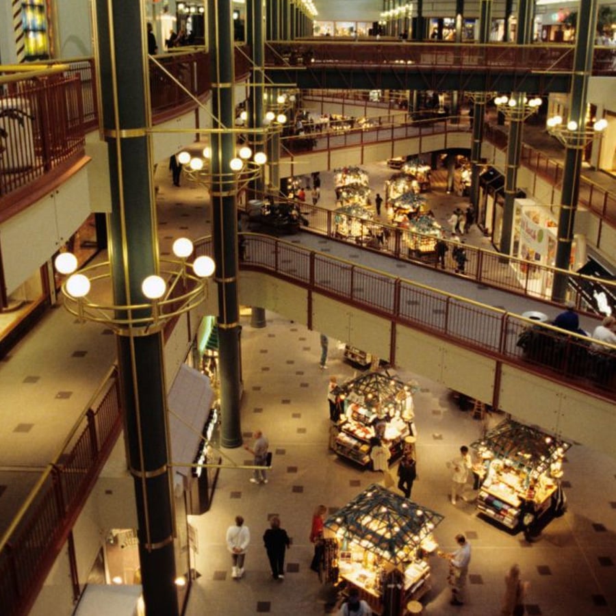 A multilevel indoor shopping mall with several kiosks on the ground floor, people walking, warm yellow lighting, and railings around the upper floors.