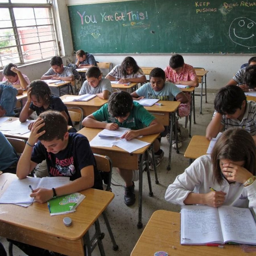 Students sit at desks, concentrating on their work in a classroom. Some have hands on their heads. A chalkboard in the background has encouraging messages like “You’ve Got This!” written on it.