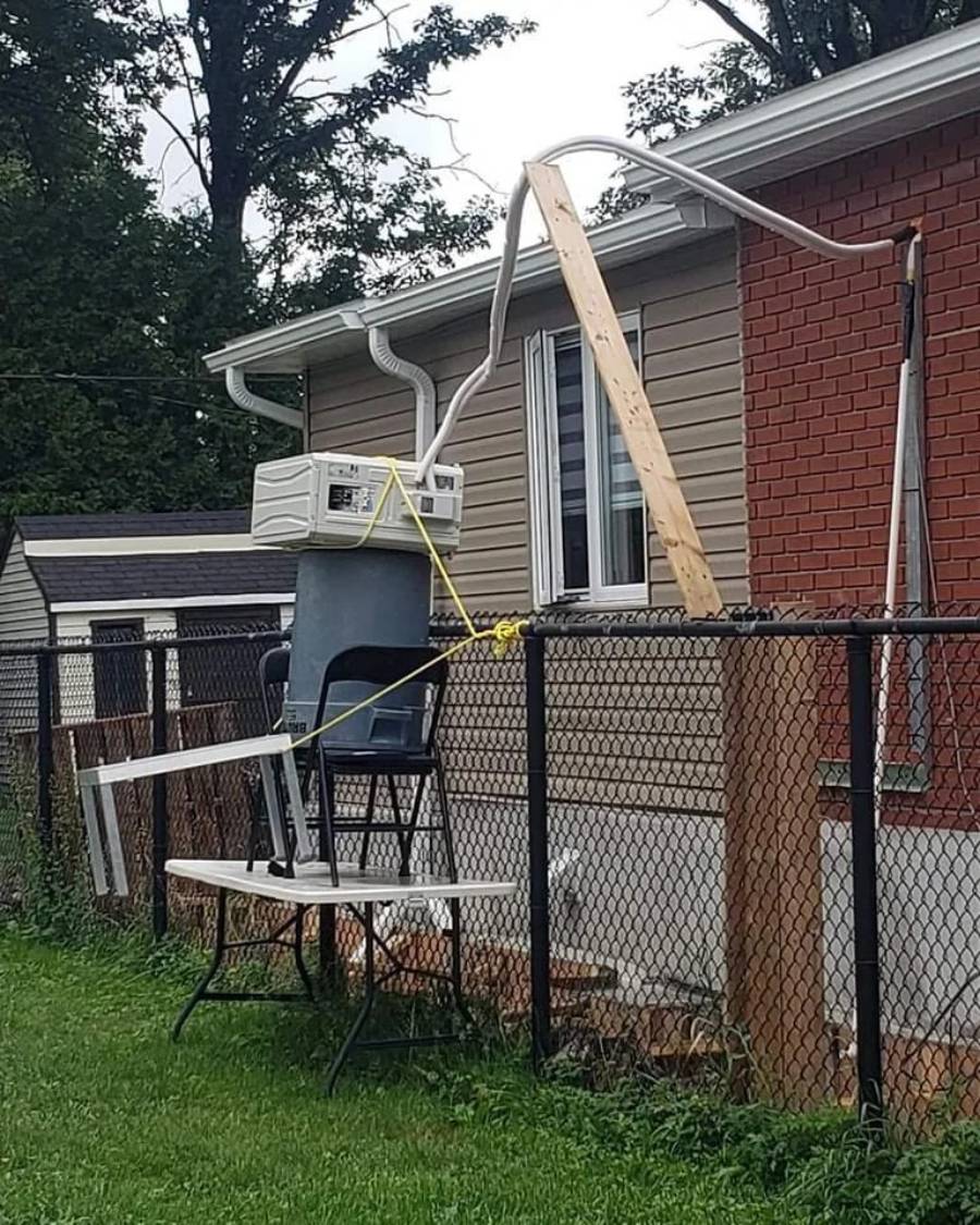 A window air conditioner is propped on a trash bin and chair, supported by wood and duct-taped to a fence, with hoses running into a house. The setup appears unstable and improvised.