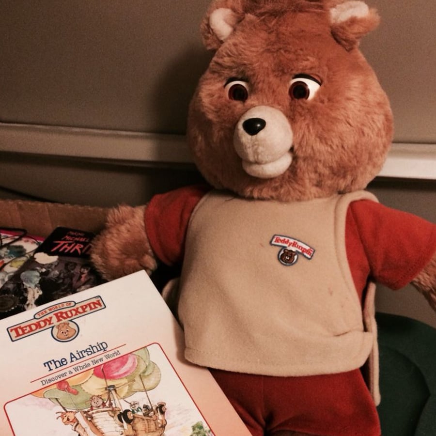 A Teddy Ruxpin bear plush toy wearing a tan vest sits next to a Teddy Ruxpin book titled “The Airship,” featuring an illustration of characters in a hot air balloon.