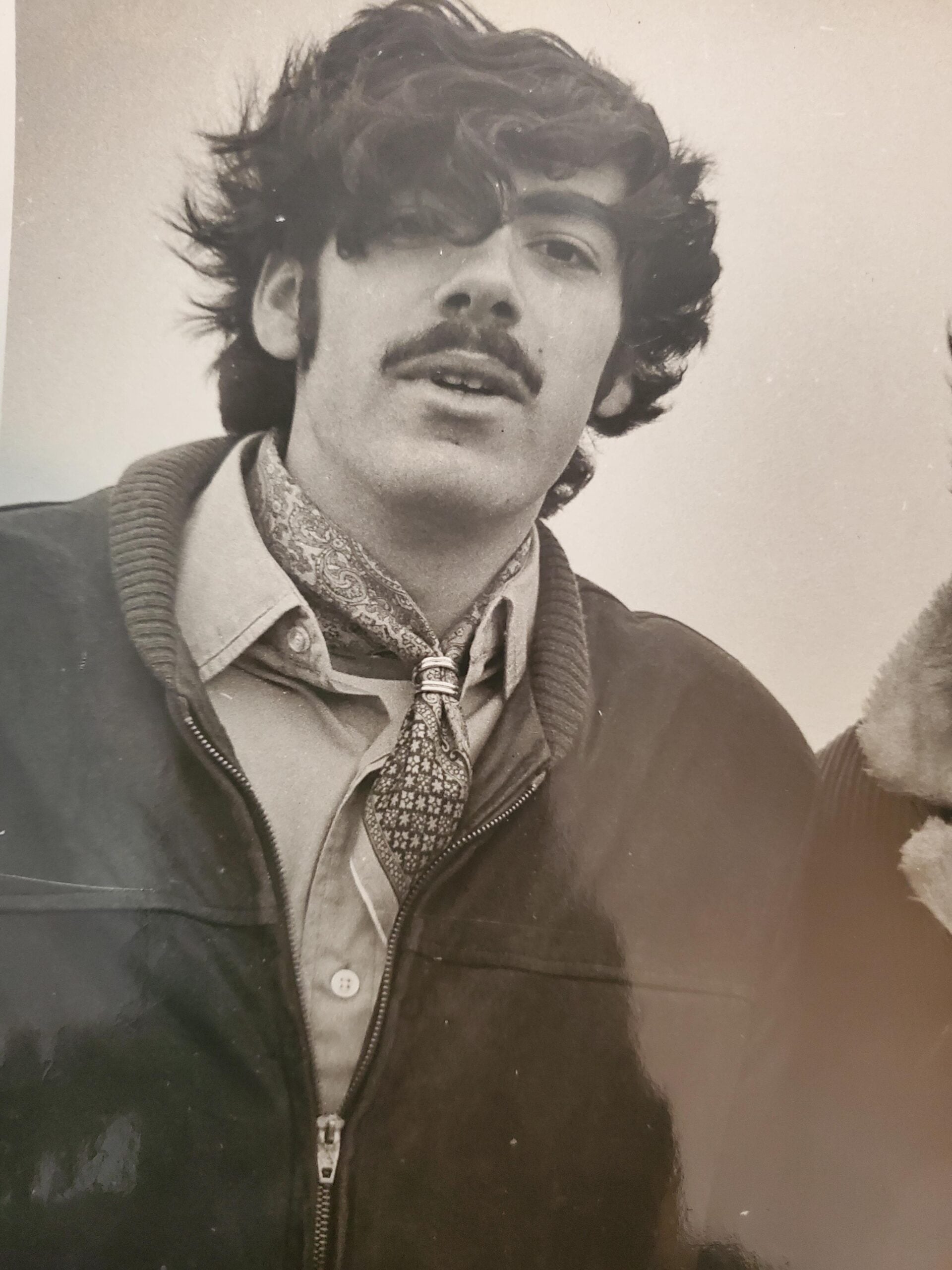 A young man with wavy hair and a mustache wears a zipped-up jacket, collared shirt, and patterned scarf tied around his neck. The black-and-white photo has a vintage feel.