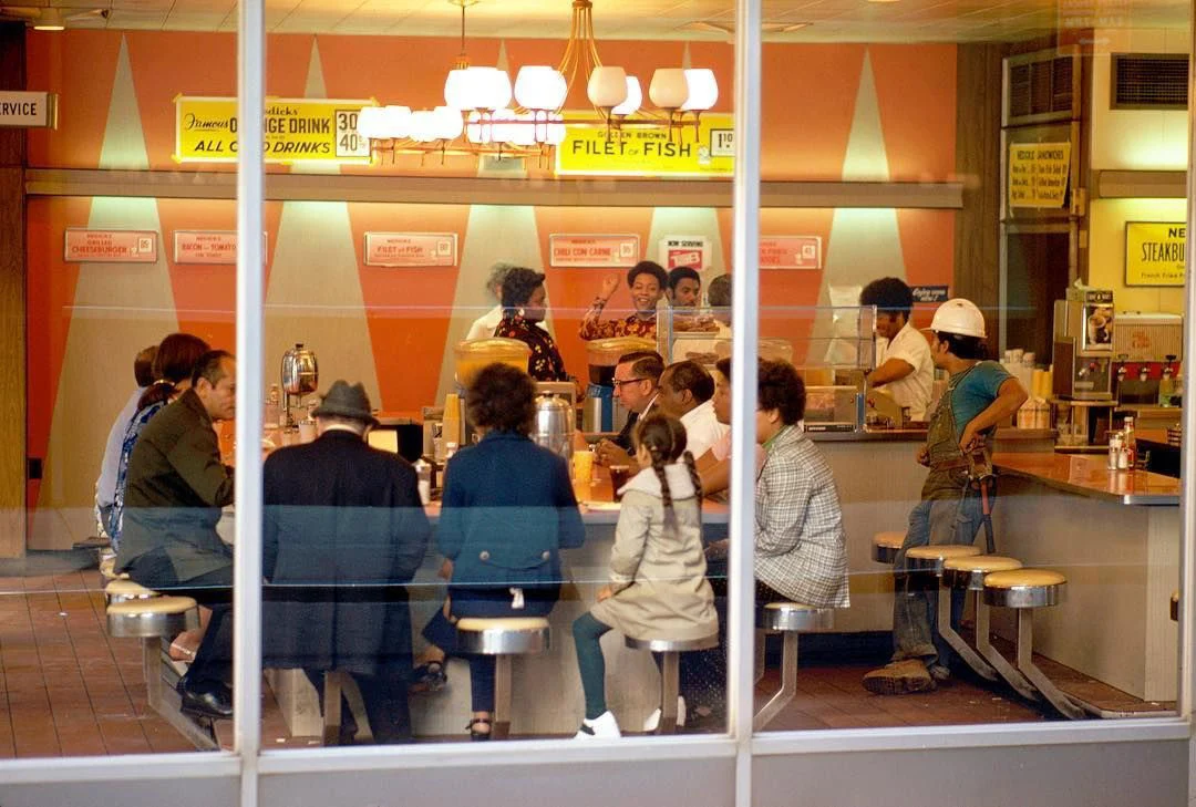 People sit at a diner counter and tables, seen through a large window, with workers and staff behind the counter. The decor is retro, with vintage signs and pastel colors.