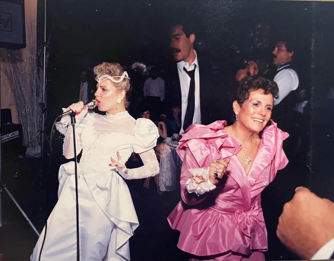 Two women in elaborate 1980s-style dresses, one white and singing into a microphone, the other pink and dancing, are at a lively party with people in formal attire in the background.