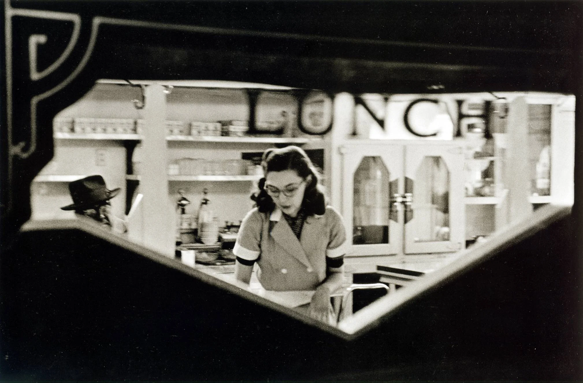 A woman in a vintage diner uniform stands behind a counter, seen through a window. Shelves and cabinets with dishes are in the background, and part of the word “LUNCH” is visible on the wall.