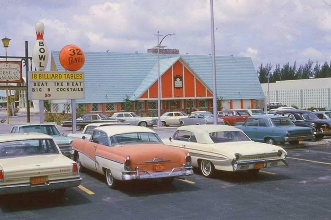 A vintage bowling alley with a retro orange and white exterior is seen behind several classic 1950s and 1960s cars parked in front. A sign advertises 32 lanes and 18 billiard tables. The sky is partly cloudy.