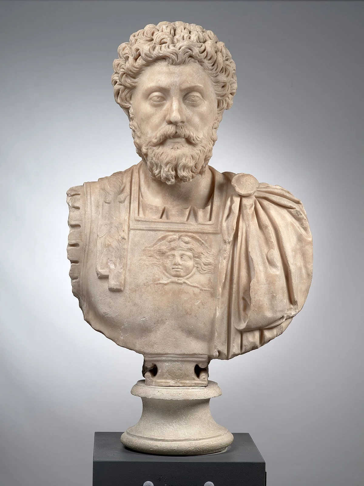 Marble bust of a bearded man with curly hair, wearing a detailed draped garment and a breastplate featuring a Medusa head, displayed on a pedestal against a plain background.