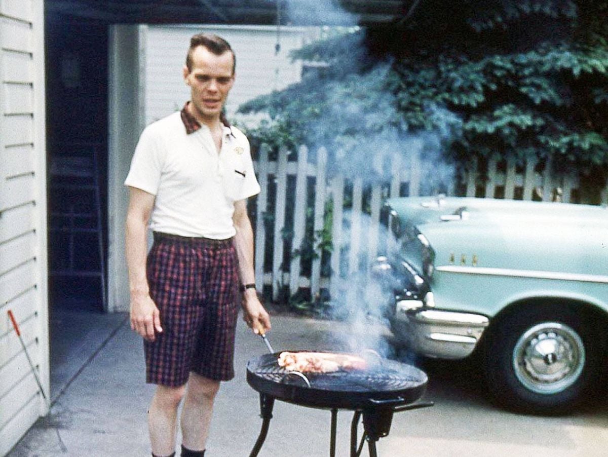 A man wearing a white shirt and plaid shorts grills meat on a charcoal BBQ in a driveway, with smoke rising. A vintage car and a white picket fence are in the background.