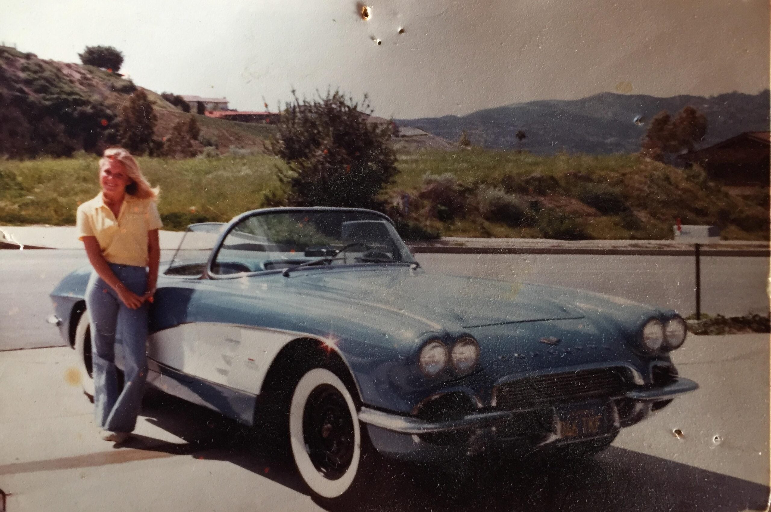 A woman in a yellow shirt and jeans stands smiling beside a blue vintage convertible car, parked on a driveway with green hills and a house in the background on a sunny day.