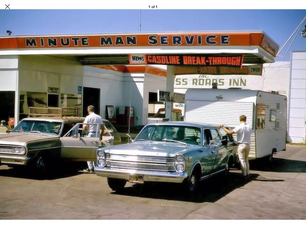 Two men stand by classic cars at a vintage gas station named “Minute Man Service,” with a trailer hitched to one car. The station has a bold red and orange sign and an old-fashioned design.