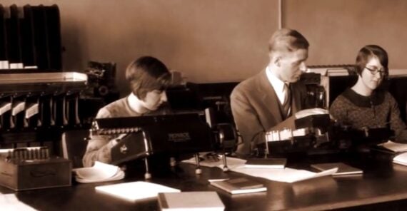 Four people sit at a desk in an old-fashioned office, working on early mechanical calculators with papers and books scattered around. The image is sepia-toned, suggesting it is from the early 20th century.