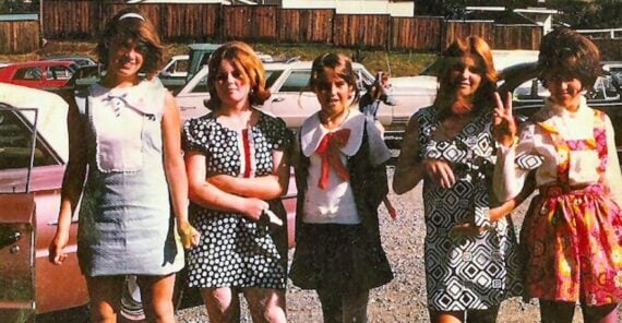 Five young women stand outdoors in front of parked cars, wearing colorful, patterned 1960s-style dresses. They pose and smile, with bright sunlight casting shadows on the gravel ground.