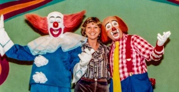 A woman smiles standing between two clowns in colorful costumes, one in blue with red hair and one in red plaid with a yellow tie, against a green and orange circus-themed background.