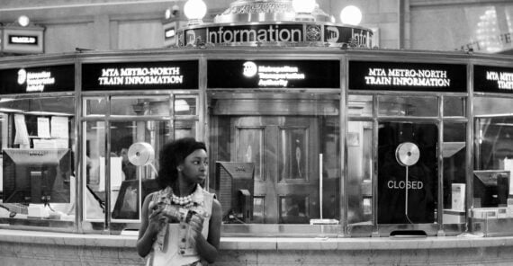 A woman stands in front of the information booth under a large clock at Grand Central Terminal, holding a drink and looking to the side. The sign reads "MTA Metro-North Train Information.