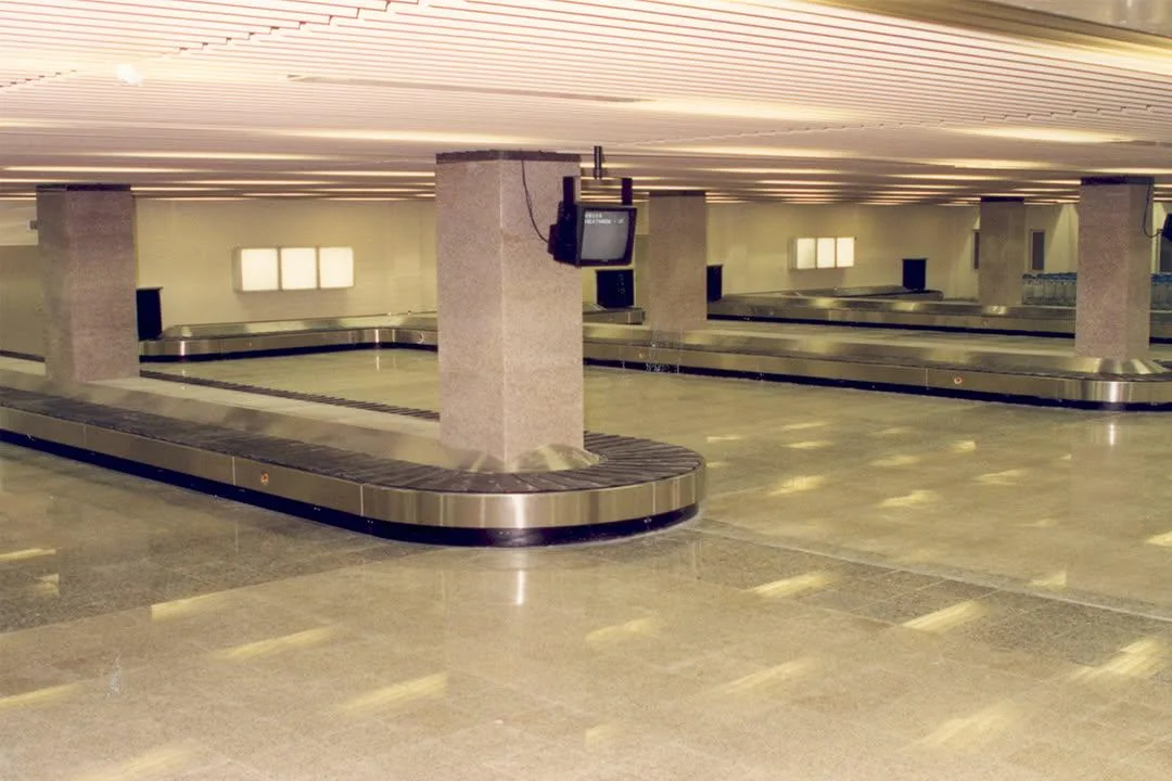 An empty airport baggage claim area with multiple conveyor belts, large pillars, a wall-mounted TV, and bright overhead lighting. No people or luggage are present.