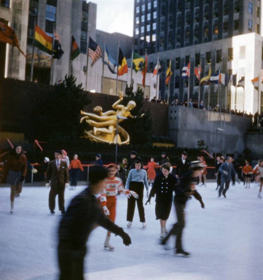 People ice skating at Rockefeller Center in New York City, with colorful flags and the golden Prometheus statue in the background. The scene is lively with skaters in winter clothing.