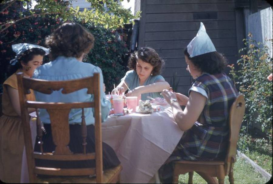 Four young women sit around a table outdoors, wearing party hats and enjoying drinks and snacks. The table is covered with a pink tablecloth, and the scene is set in a sunny backyard with greenery and flowers.