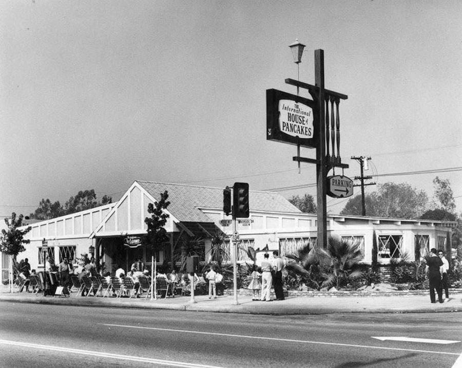 Black and white photo of the original International House of Pancakes restaurant, with people gathered outside and a large sign reading "International House of Pancakes" and "Parking" near a street intersection.