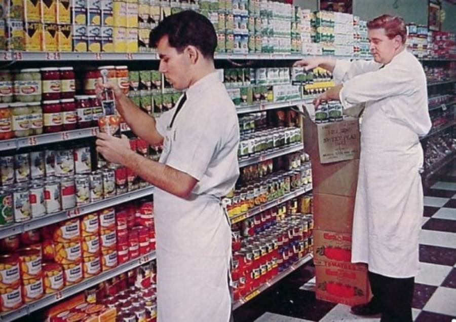 Two men wearing white aprons are stocking canned goods on supermarket shelves. One arranges cans on a lower shelf while the other unpacks boxes, with shelves filled with various canned products around them.