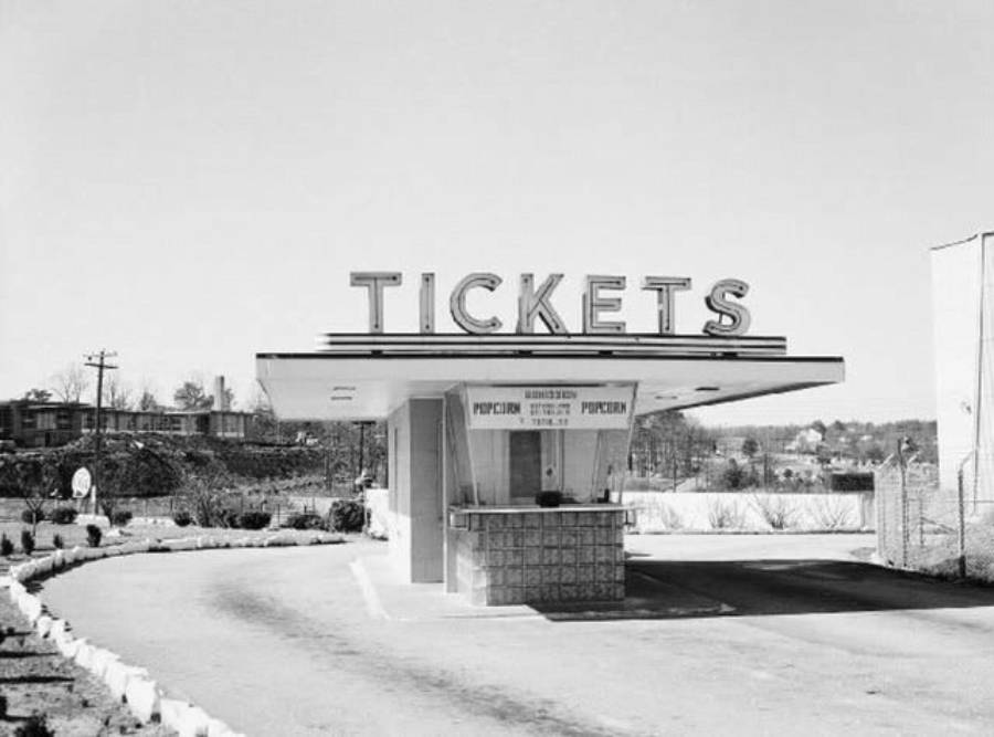 A vintage black-and-white photo of a small, standalone ticket booth with a large "TICKETS" sign on top, located in an empty drive-in lot with landscaping and distant buildings in the background.