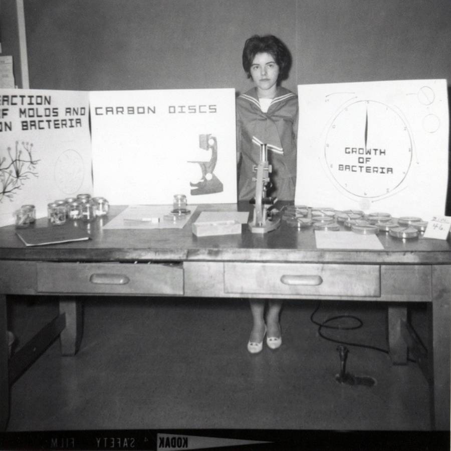 A young woman stands behind a table displaying science fair projects with posters about molds, bacteria, and carbon discs, petri dishes, and a microscope.