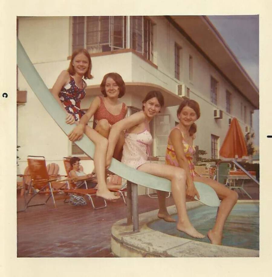 Four girls in swimsuits sit on a pool slide, smiling at the camera. They are outdoors by a pool, with a hotel building, orange chairs, and umbrellas visible in the background. The photo has a vintage feel.