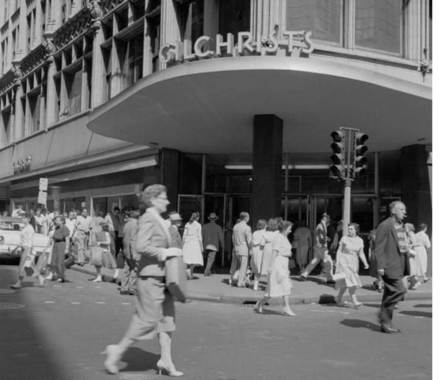 Black and white photo of people crossing a busy city street in front of a department store with a large "Gilchrist's" sign above the entrance. The scene appears to be from the mid-20th century.