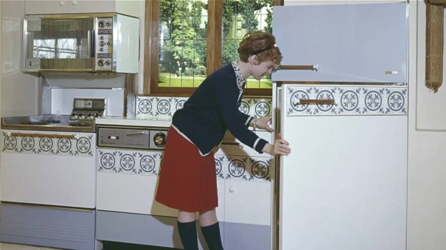 A woman in vintage clothing opens a retro refrigerator in a kitchen with patterned tiles, with appliances and a window showing trees in the background.