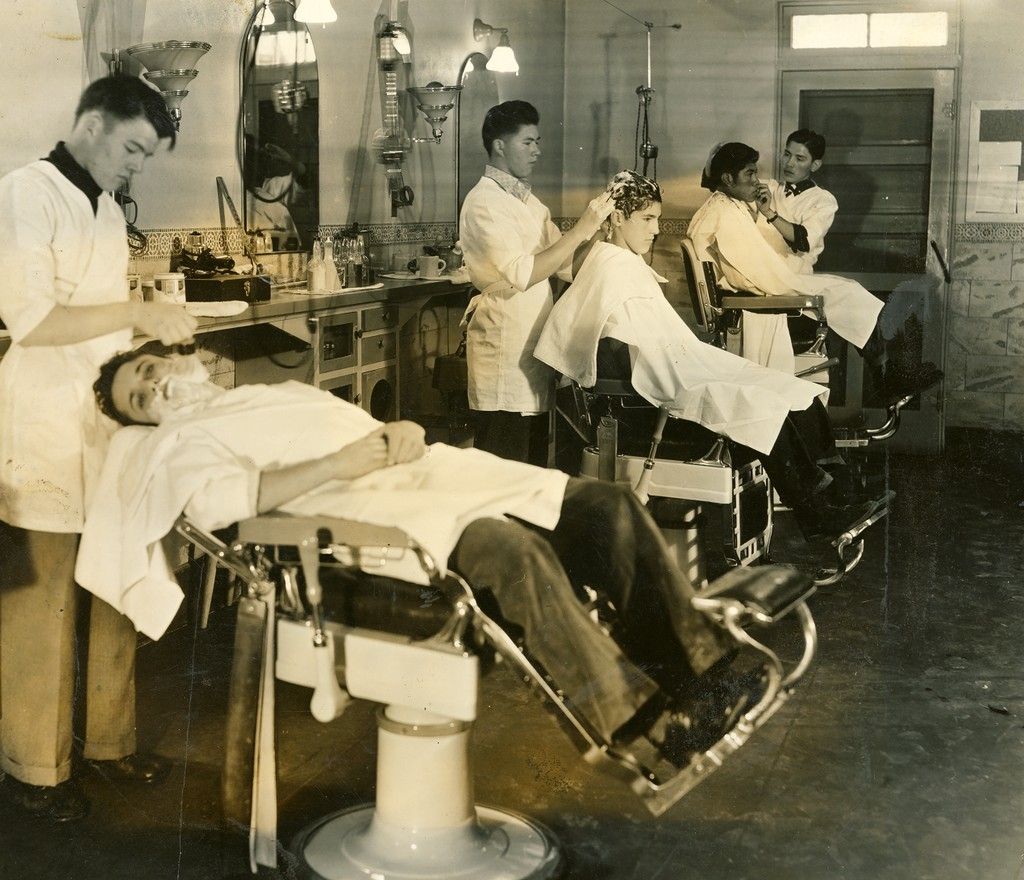 Three men in barber chairs receive haircuts and shaves from barbers in white coats in a vintage barbershop, with mirrors and shelves lining the wall.