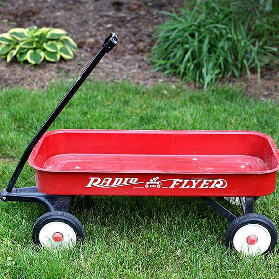 A classic red Radio Flyer wagon with a black handle sits on green grass, with plants and soil in the background.