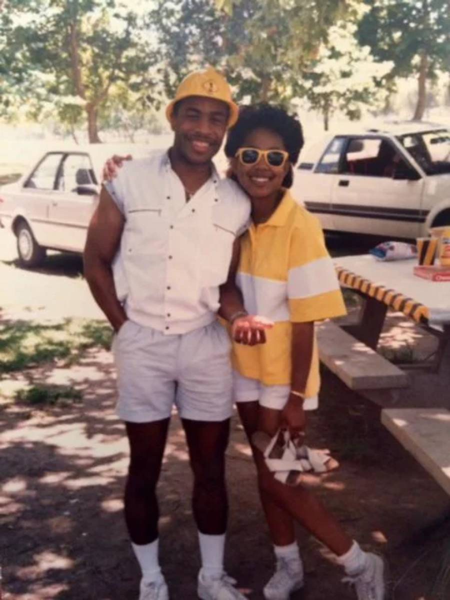 Two people in 80s-style summer outfits smile and pose outdoors by a picnic table. Parked cars and trees are in the background. The woman holds her sunglasses, and both look happy and relaxed.