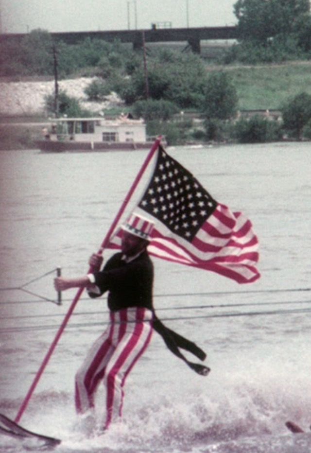 A person dressed in a patriotic Uncle Sam costume water-skis on a river while holding a large American flag, with a boat and greenery visible in the background.