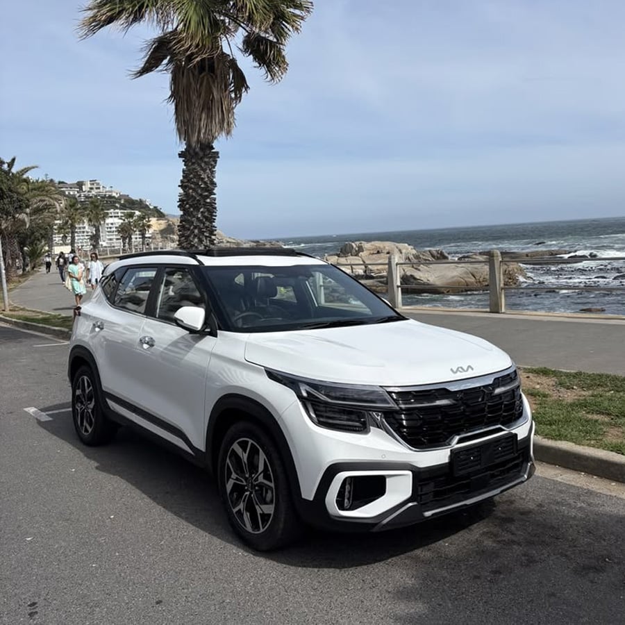 A white Kia SUV is parked on a coastal road near palm trees, with people walking and the ocean visible in the background.