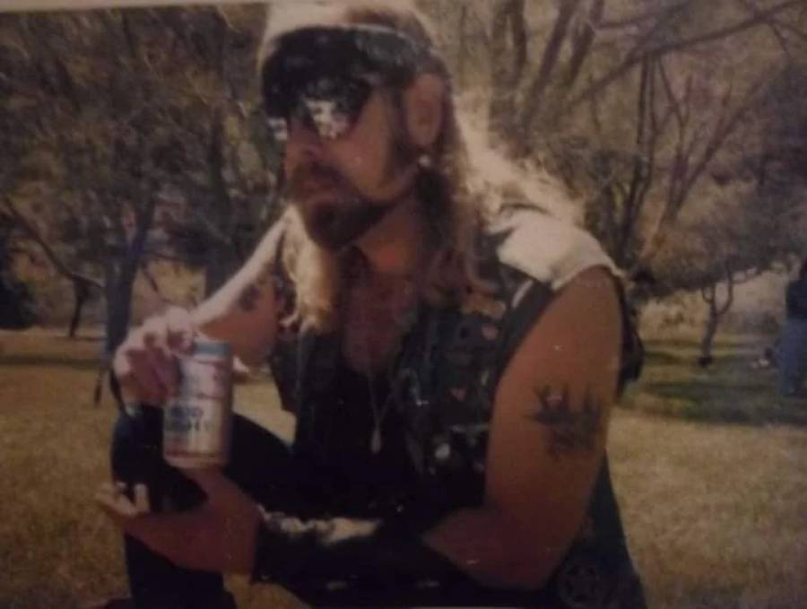 A person with long hair, sunglasses, and a bandana kneels outdoors, holding a can. They wear a sleeveless vest and have visible tattoos on their arms. The background features trees and sunlight.