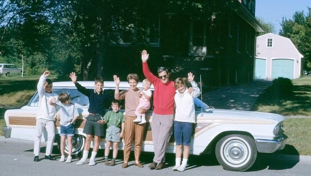 A family of seven, including four children, two adults, and a toddler, stand smiling in front of a vintage white station wagon with wood paneling, waving at the camera on a sunny day.