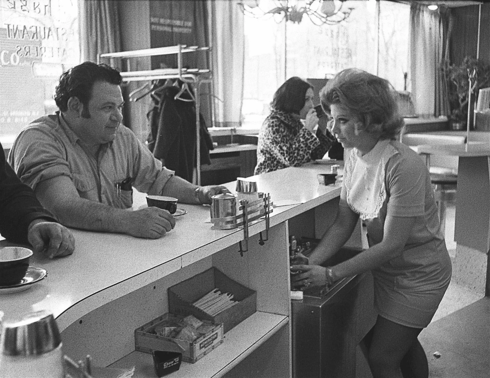 A man sits at a diner counter with a coffee cup while a woman in a short dress stands behind the counter, preparing something. Other patrons sit in the background.