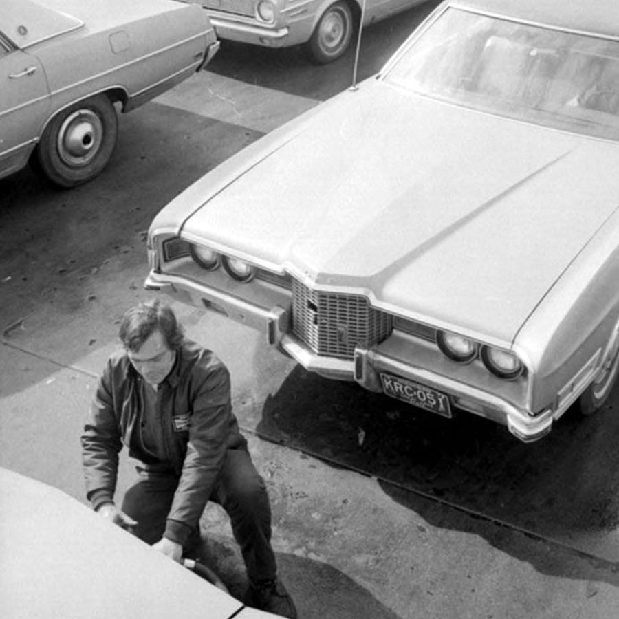 A person wearing glasses and a jacket is bending over near the hood of a car in a parking lot with several vintage cars visible around. The image is in black and white.