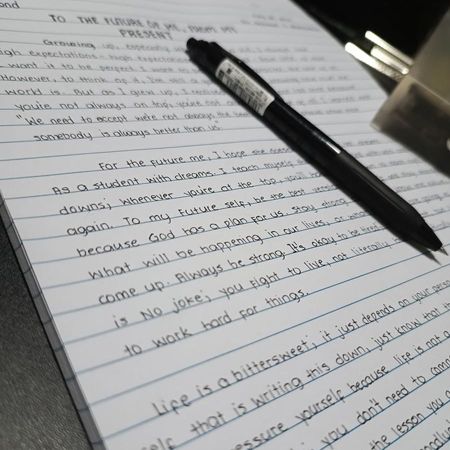 A black pen rests on lined notebook paper filled with neatly handwritten text in English. The writing discusses future aspirations and motivation. Part of a metal clip is visible in the corner.