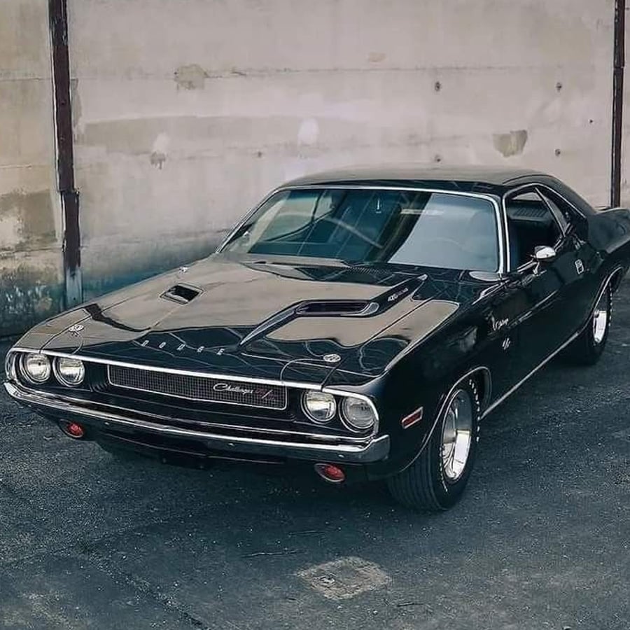 A black classic Dodge Challenger muscle car is parked on a paved surface in front of a weathered concrete wall.