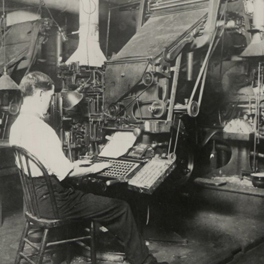 A man in a white shirt and suspenders operates a vintage typesetting machine in an old industrial print shop filled with large mechanical equipment.