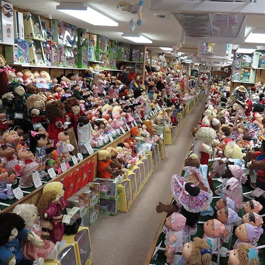 Shelves and tables filled with hundreds of Cabbage Patch Kids dolls in a brightly lit store, with dolls of various sizes, outfits, and hair colors arranged neatly throughout the space.