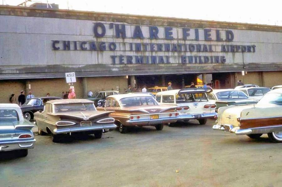 Vintage cars parked in front of the O’Hare Field Chicago International Airport Terminal Building, with people walking near the entrance and the airport sign visible above.