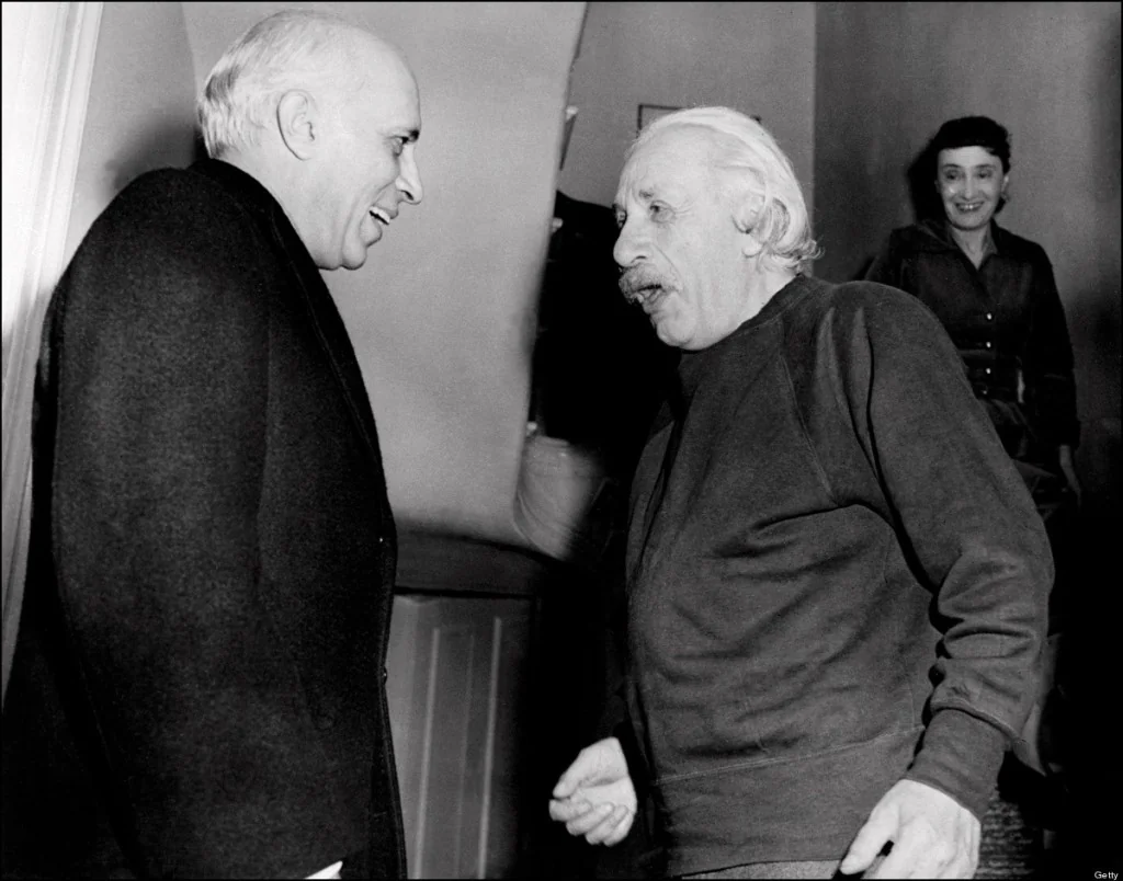 Two older men stand and talk animatedly indoors while a smiling woman watches from the stairs in the background. The scene appears candid and friendly, with all three subjects engaged and expressive.