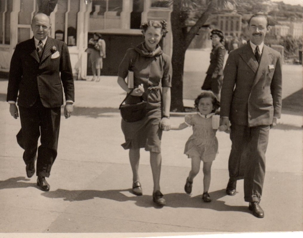 Three adults and a young girl walk together on a sunny sidewalk. The girl holds hands with two adults and smiles. Two men wear suits, and the woman wears a dress. A uniformed person and buildings are visible in the background.