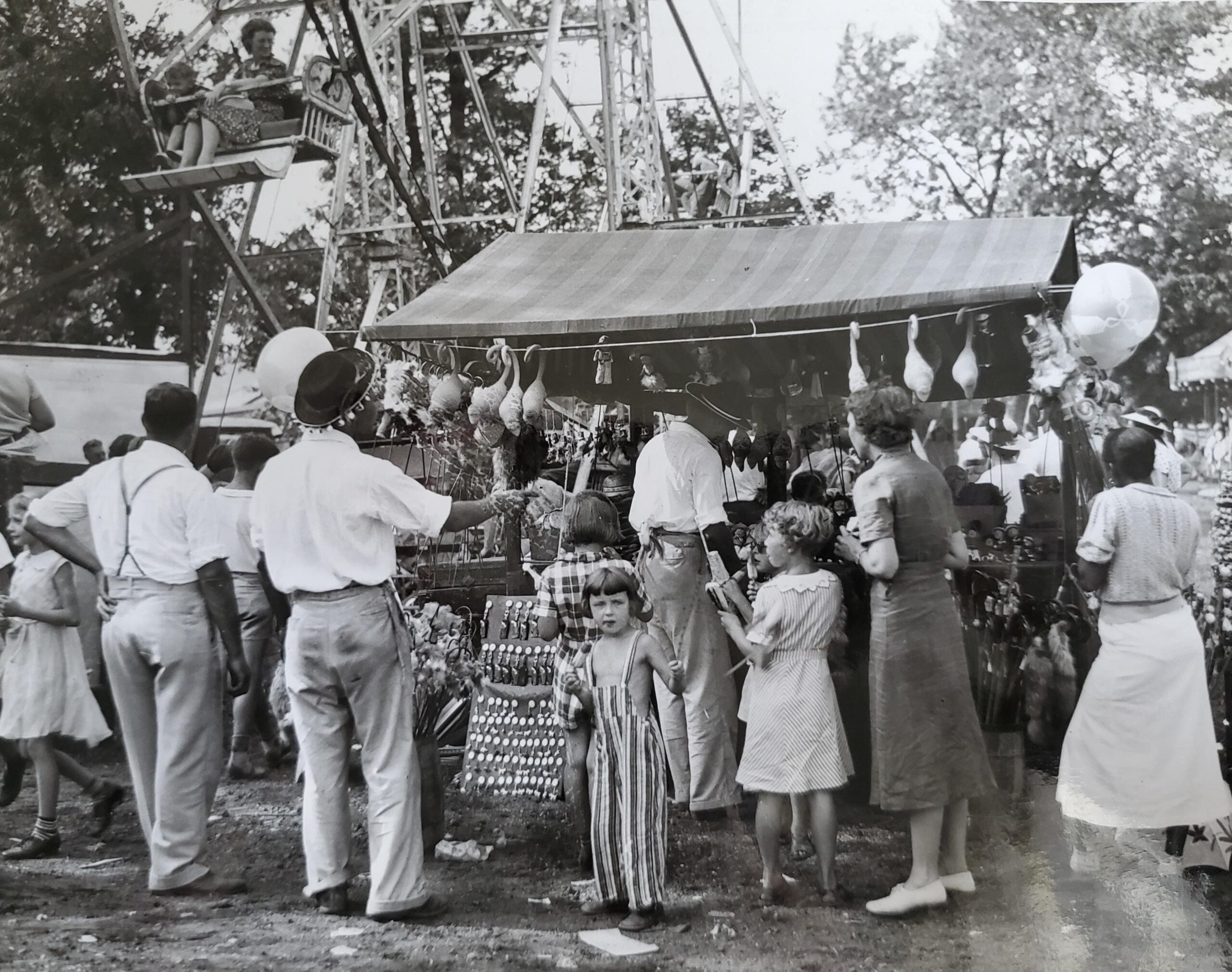 A black and white photo of a busy fairground with people gathered around a booth selling toys and prizes. Children and adults stand near the stall, while a ferris wheel operates in the background among tall trees.
