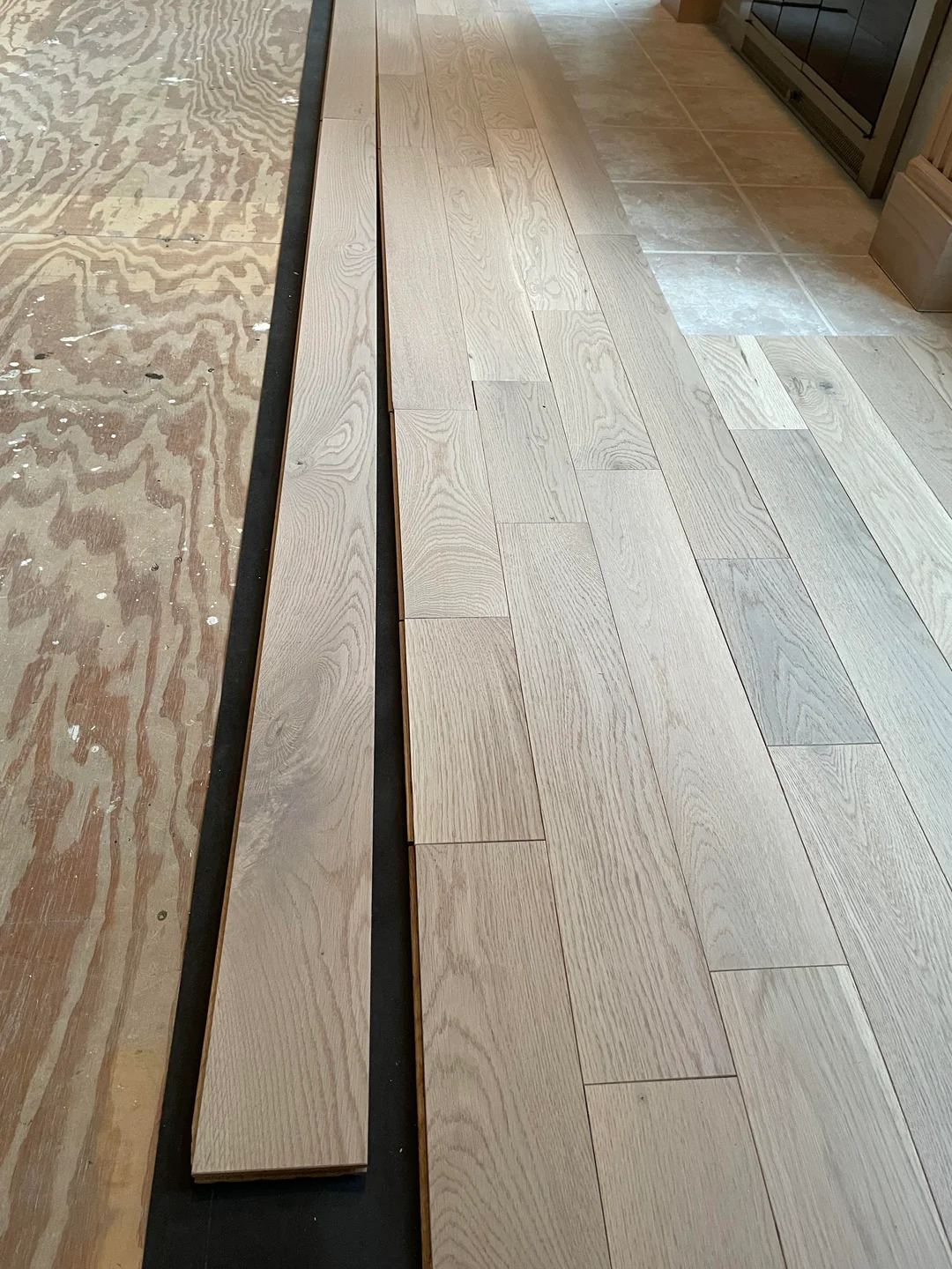 Light wood flooring planks being installed next to plywood subfloor and beige tile, with a black underlayment separating the new planks from the unfinished area.