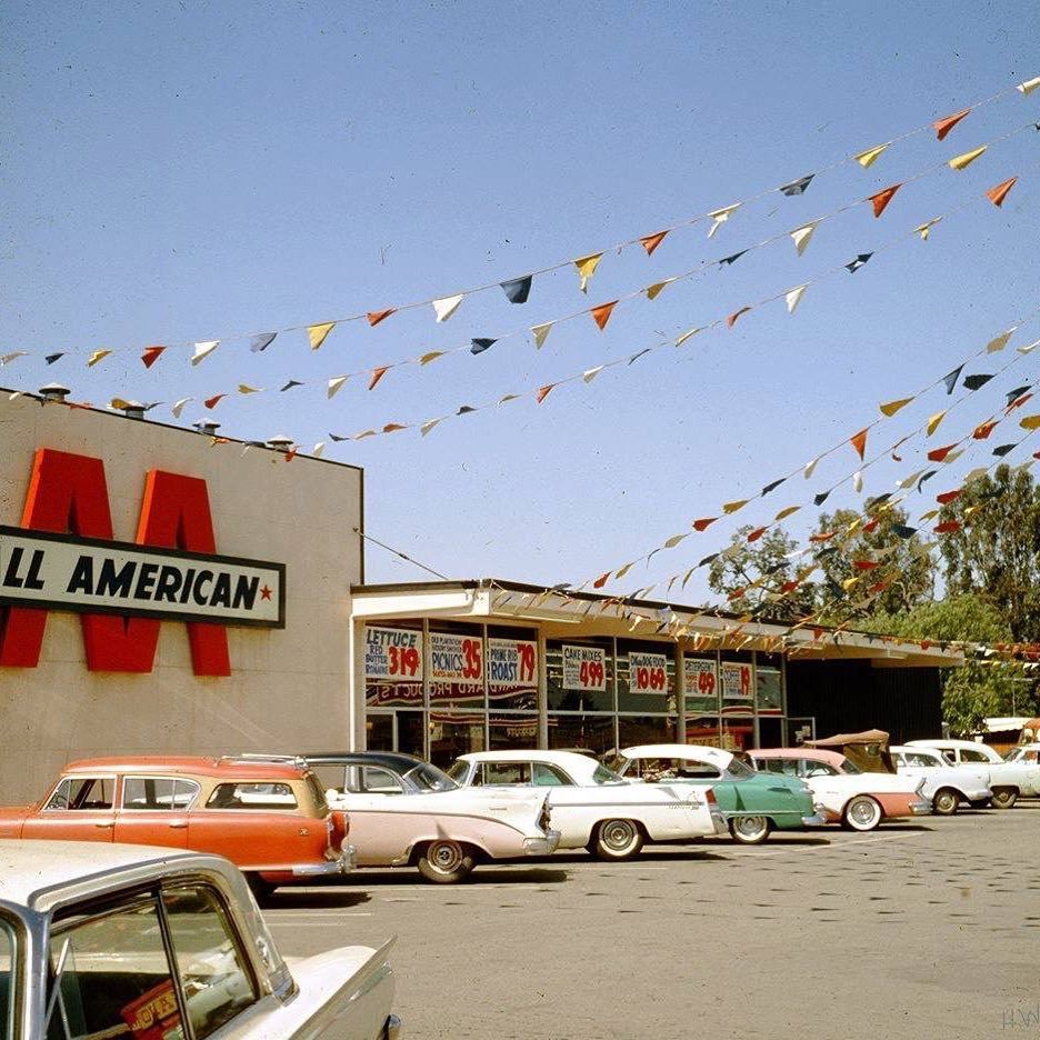 A 1950s supermarket parking lot with colorful pennant flags overhead, vintage cars parked in front, and a store sign reading “ALL AMERICAN.” Storefront windows display large posters for groceries.