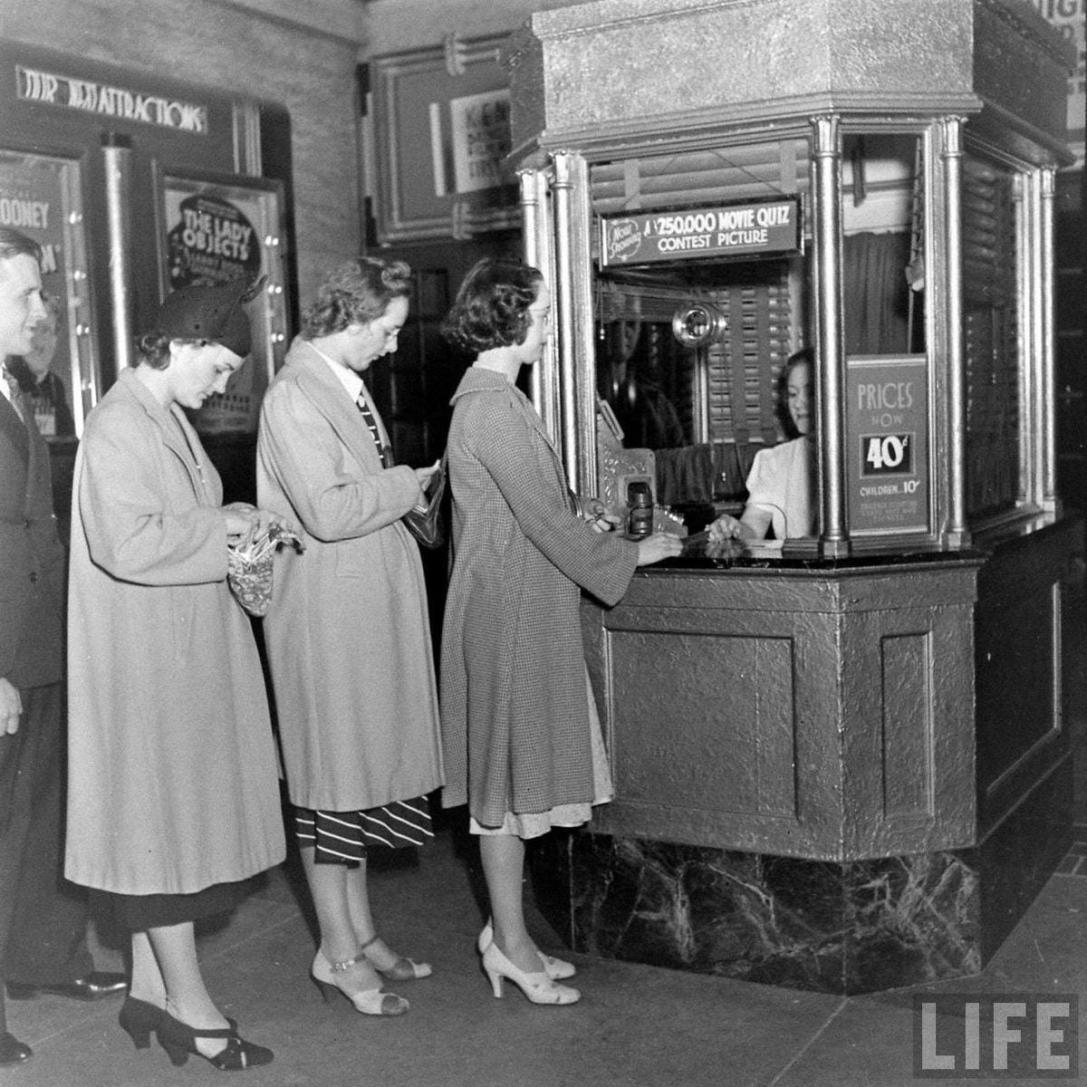 Three women stand in line at a movie theater ticket booth, speaking with a cashier behind glass. A man stands behind them. Signs display ticket prices and a movie quiz contest. The setting appears vintage, likely mid-20th century.